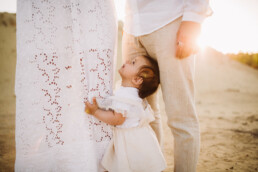 Ein kleines Mädchen hält sich, im Rahmen eines Fotoshooting bei Sonnenuntergang, am Kleid ihrer Mutter fest.
