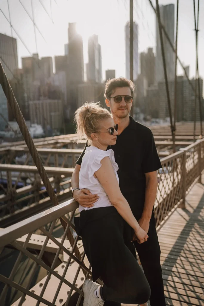 Ein Paar mit Sonnenbrille steht eng beieinander auf einer Brücke, mit den Wolkenkratzern der Stadt und dem Sonnenlicht im Hintergrund. Die entspannte, liebevolle Pose, aufgenommen von Nadja Morales, deutsche Fotografin in den USA, unterstreicht ihre Verbundenheit.