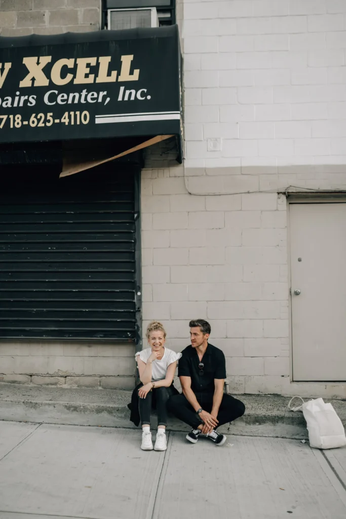 Zwei Personen sitzen während eines Fotoshootings in New York auf dem Bürgersteig vor einem geschlossenen Geschäft mit einem schwarzen Schild. Der eine lacht mit der Hand im Gesicht, der andere lächelt, während in der Nähe eine weiße Tasche auf dem Boden liegt.