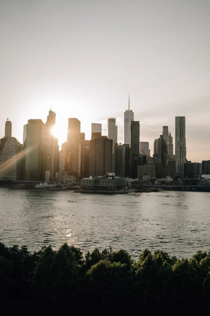Die Sonne geht hinter der Skyline von New York mit ihren hohen Wolkenkratzern unter und spiegelt sich auf dem Fluss wider. Grüne Bäume säumen das Flussufer, wo sich eine deutsche Fotografin in den USA auf ein Fotoshooting in der Abenddämmerung vorbereitet.