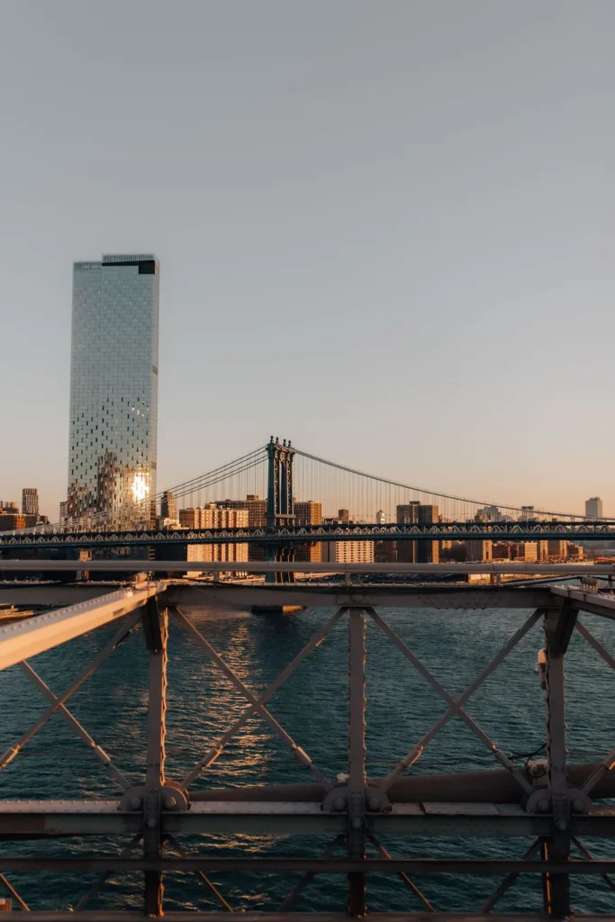 Ein Blick auf die Manhattan Bridge und die New Yorker Skyline mit einem hohen Glasgebäude bei Sonnenuntergang, aufgenommen hinter dem Metallgeländer einer anderen Brücke über den East River von einer deutschen Fotografin in den USA während eines Fotoshootings.